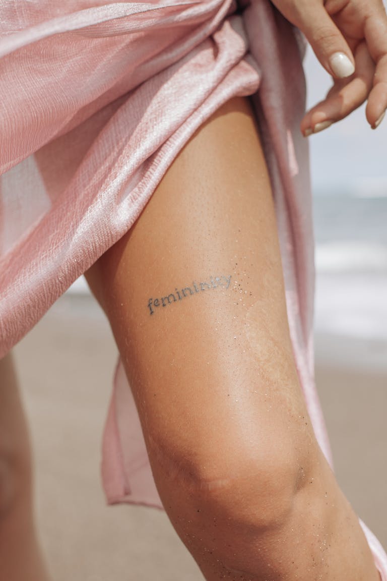 An artistic close-up of a woman's leg displaying a 'femininity' tattoo, captured on a sandy beach.
