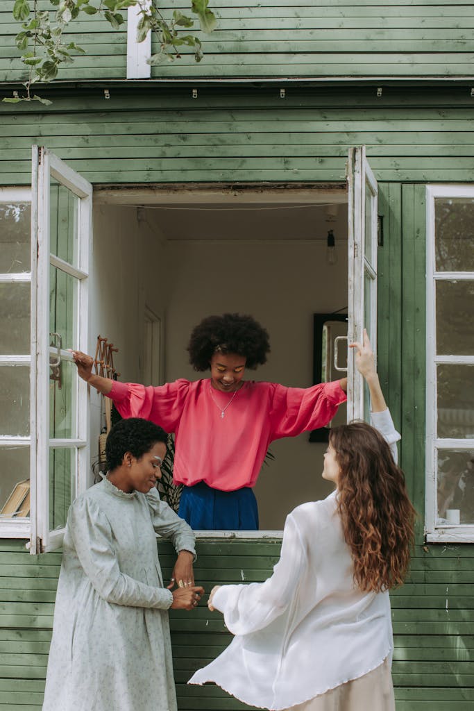 Three diverse women enjoying a cheerful moment by a rustic green cottage window in daylight.