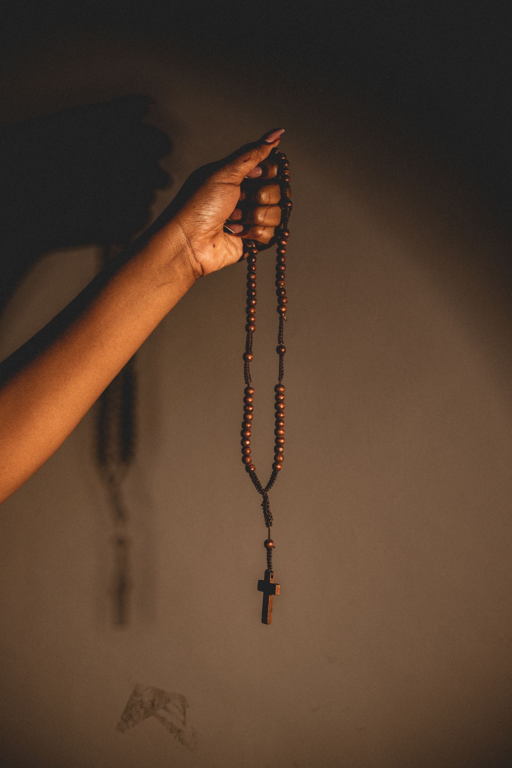 A hand gently holds wooden rosary beads with a cross, casting a shadow on the wall.