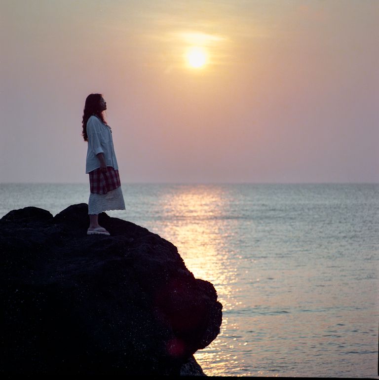 A woman stands on a rocky shore in Quang Ngai, Vietnam, watching a serene sunset.